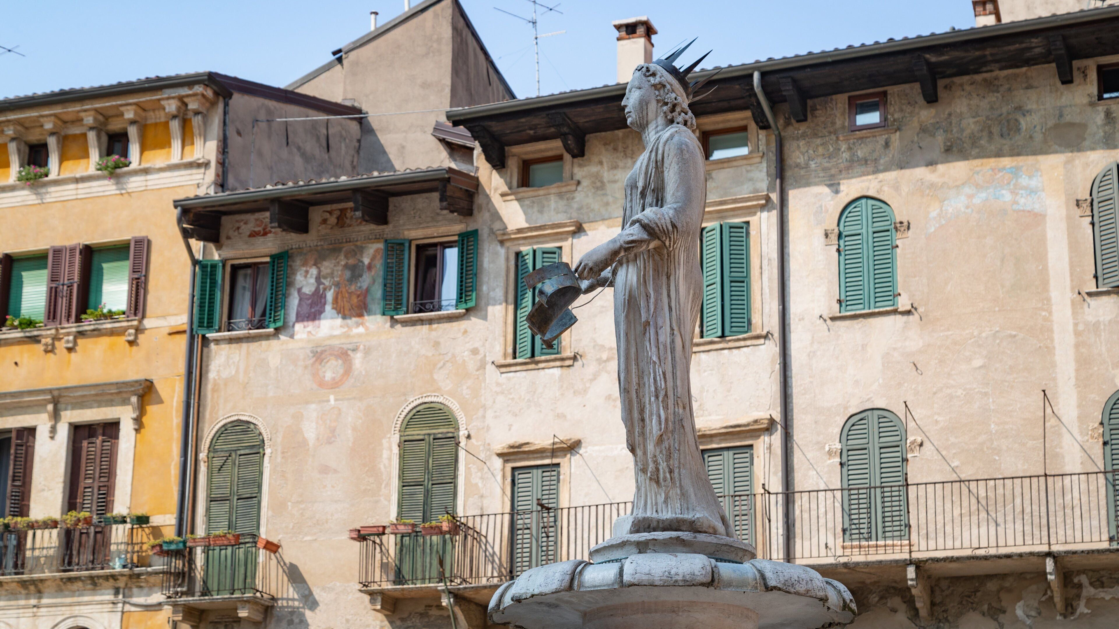 A statue of a woman holding a bucket, standing on a pedestal in front of a building with green shutters and a mural on the wall.