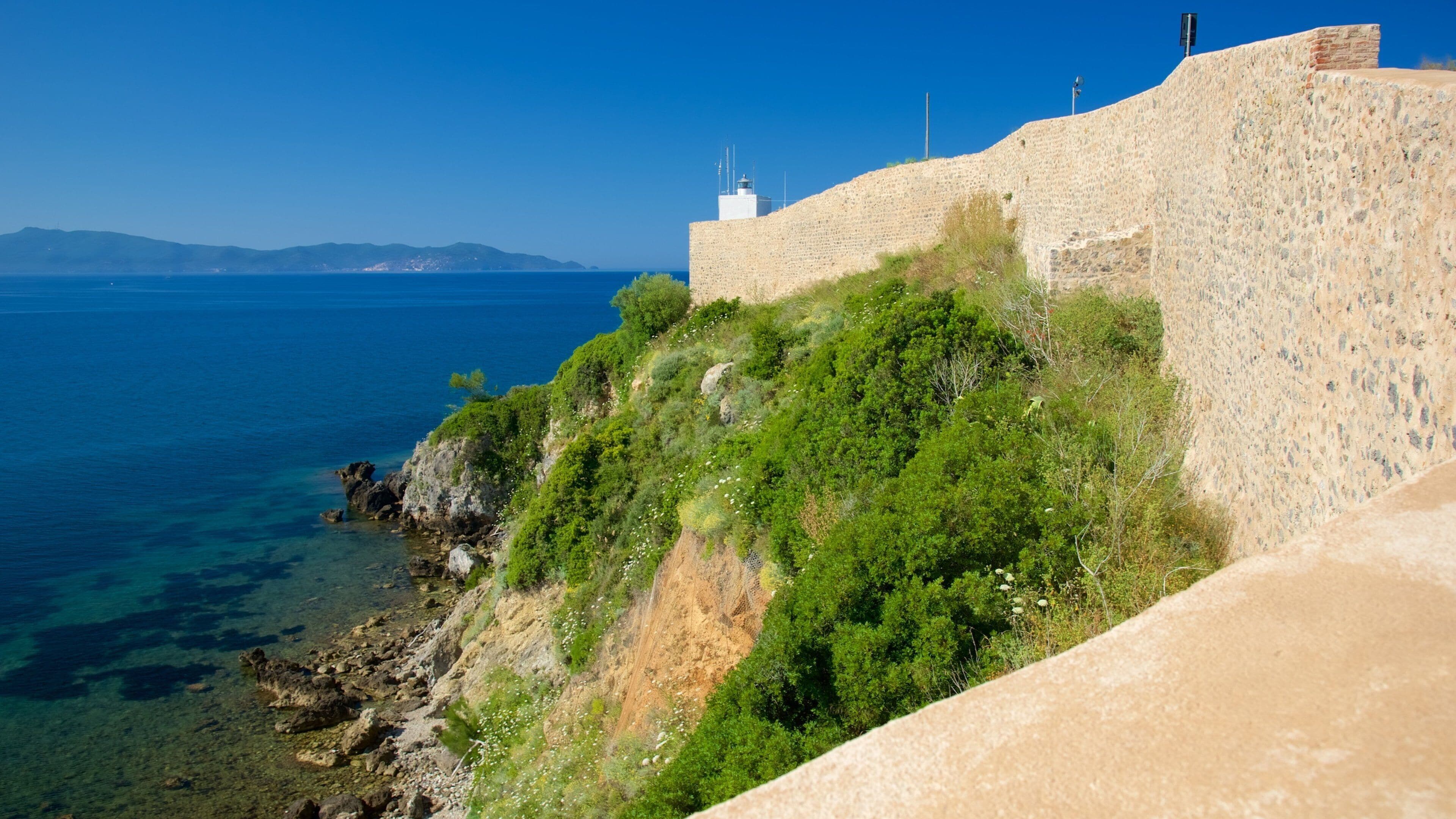 A coastal fortification with a lighthouse, clear blue sea, and distant mountains.