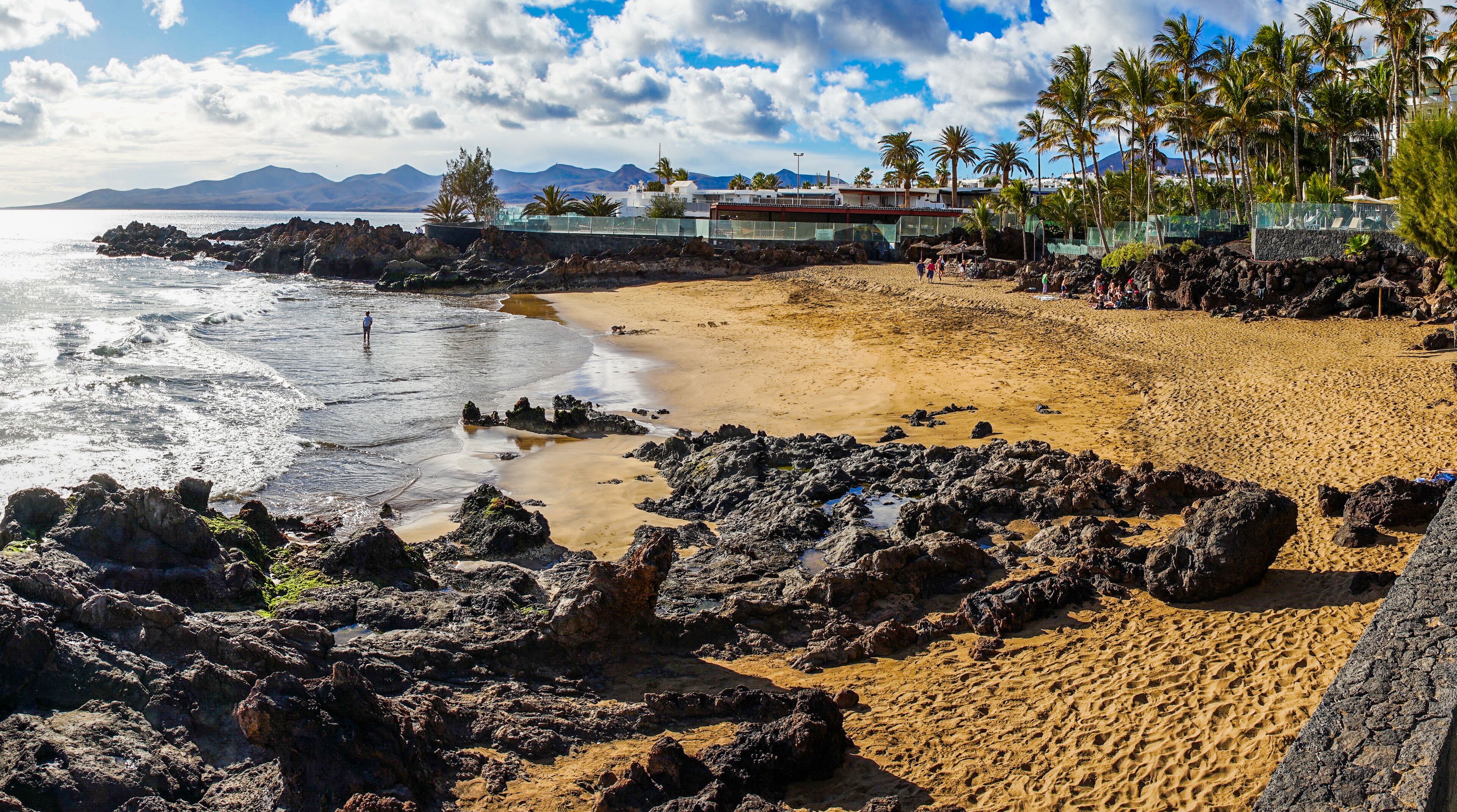 A beach with rocky foreground, sandy shore, palm trees, and distant mountains.