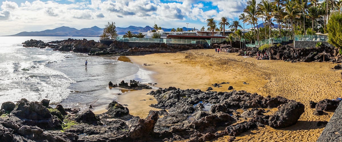 A beach with rocky foreground, sandy shore, palm trees, and distant mountains.
