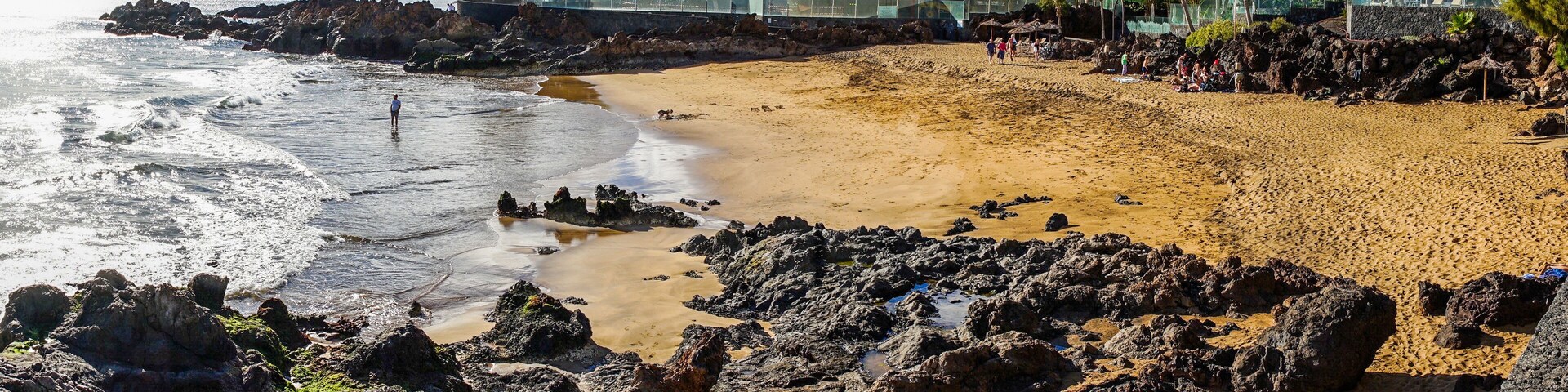 Una playa con primer plano rocoso, orilla arenosa, palmeras y montañas a lo lejos.