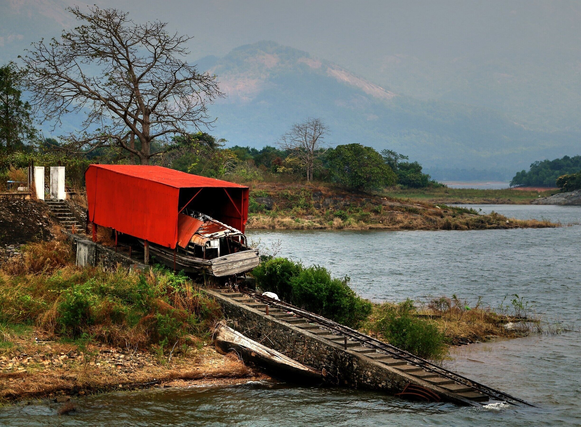 A red covered train car on a railway track beside a river, with mountains in the background.