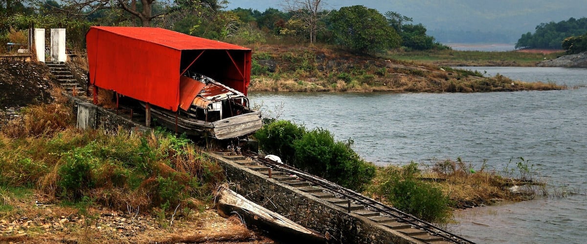 A red covered train car on a railway track beside a river, with mountains in the background.