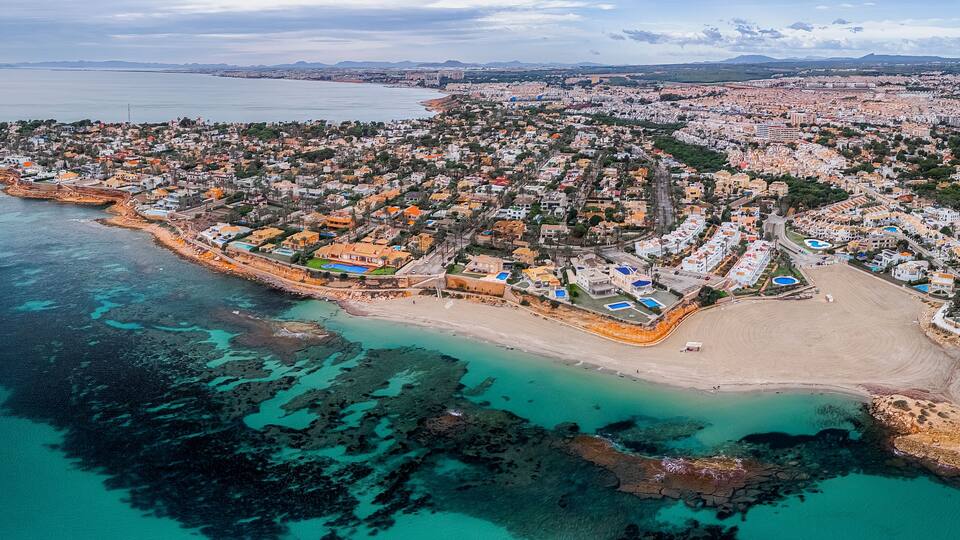 An aerial view of a coastal town with a beach, clear turquoise waters, and a mix of residential and commercial buildings.