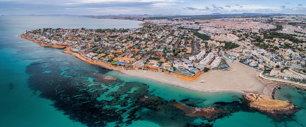 An aerial view of a coastal town with a beach, clear turquoise waters, and a mix of residential and commercial buildings.
