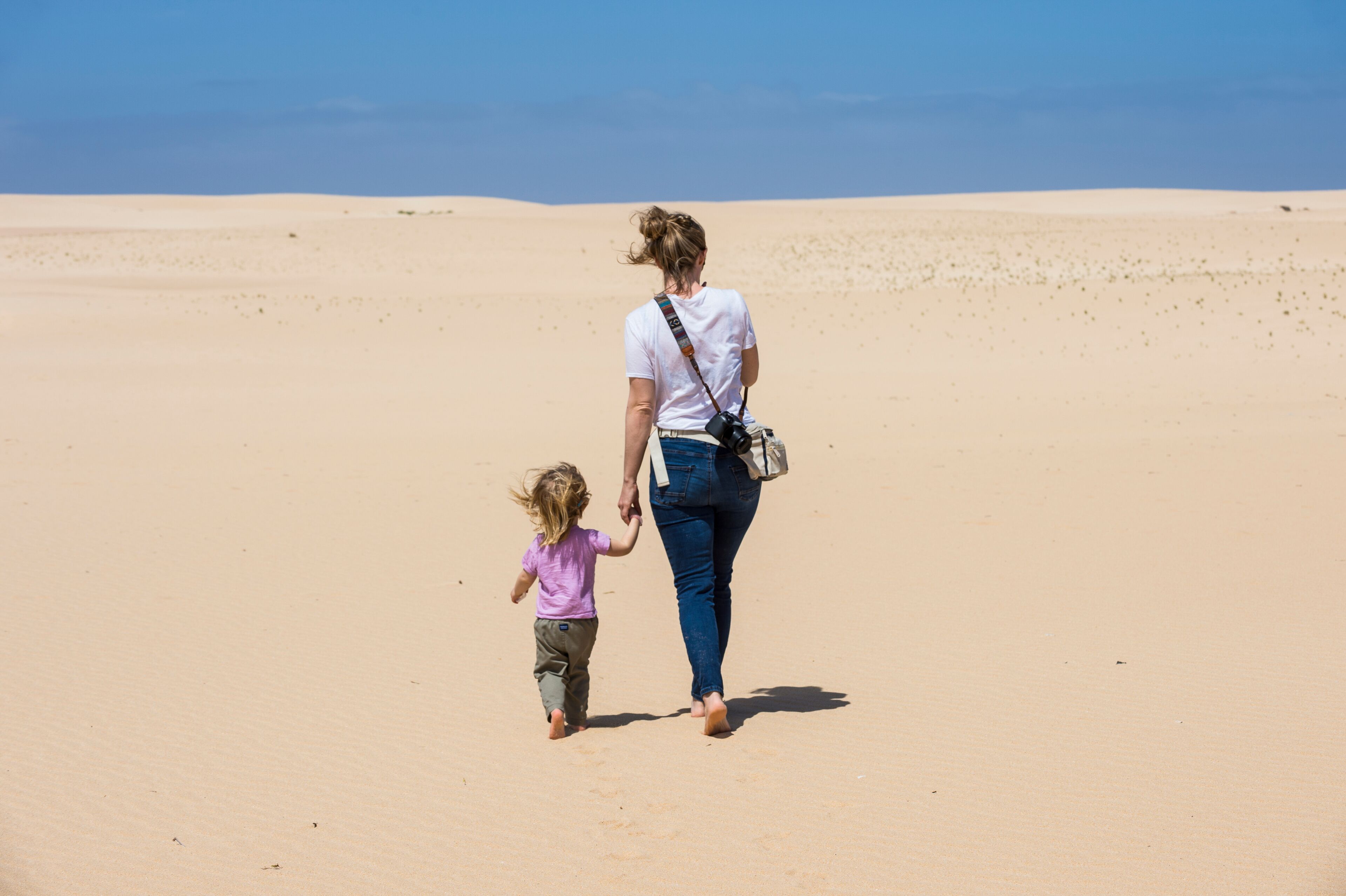 Uma mulher e uma criança caminham em uma praia arenosa sob um céu azul límpido.