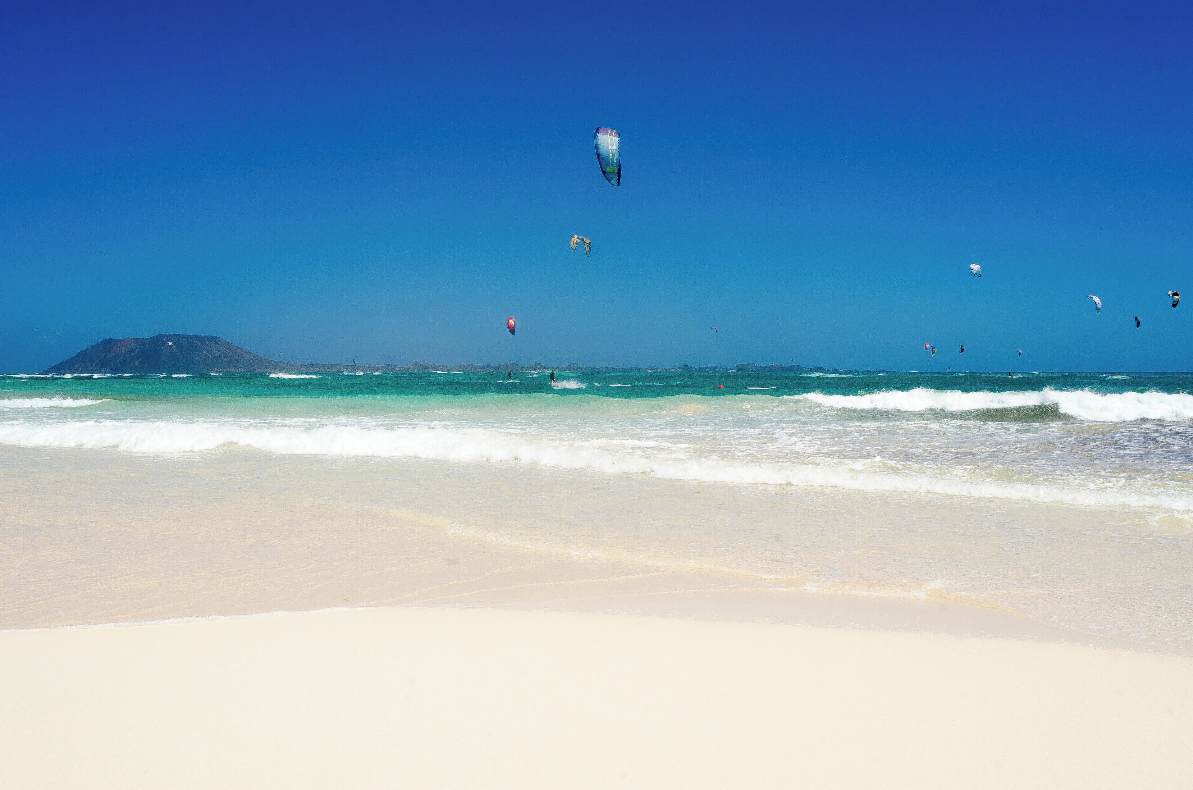 A beach with clear blue water, waves, and several kites flying in the sky.