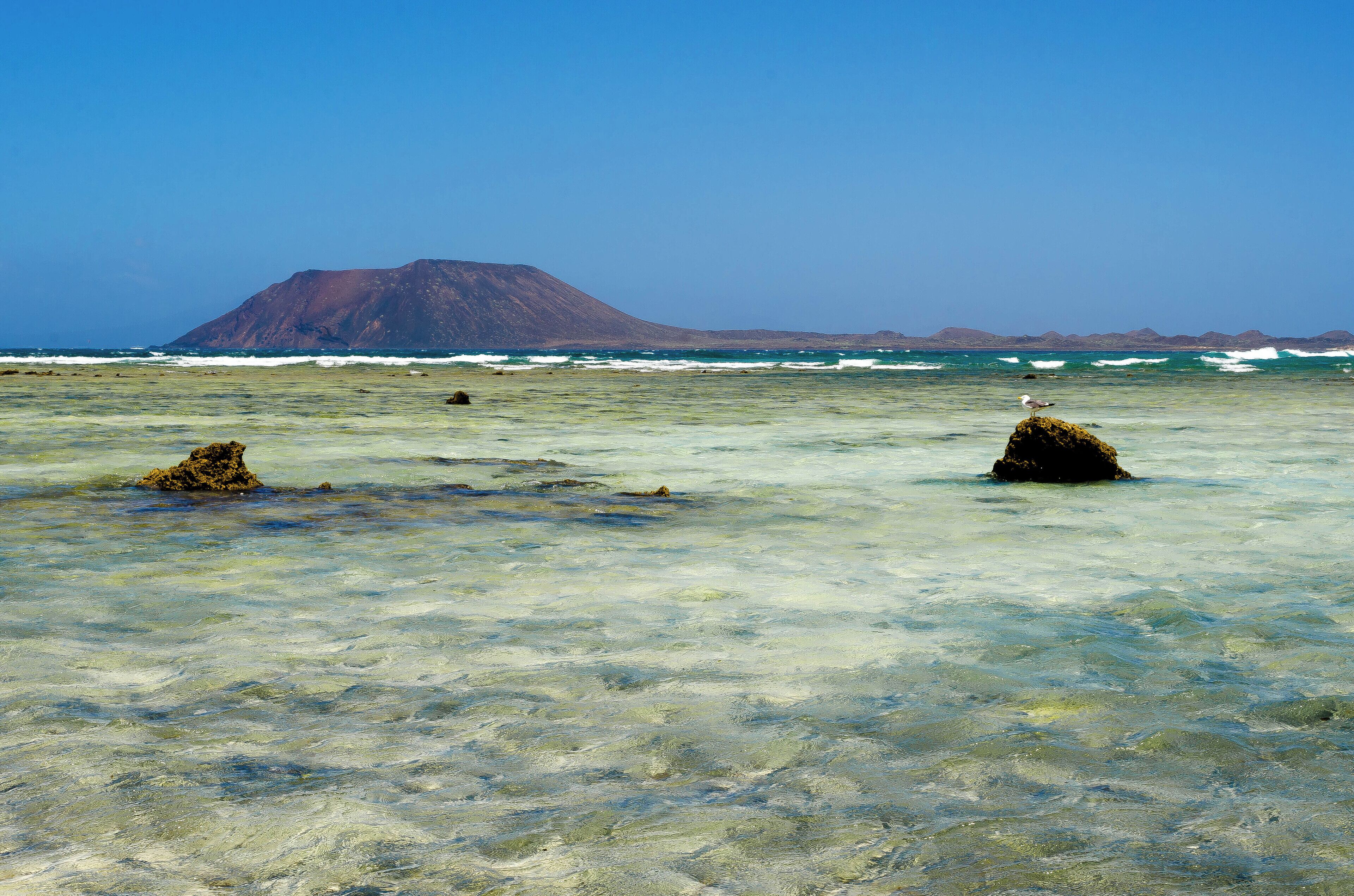 A beach with clear shallow waters, rocky outcrops, and distant mountains.