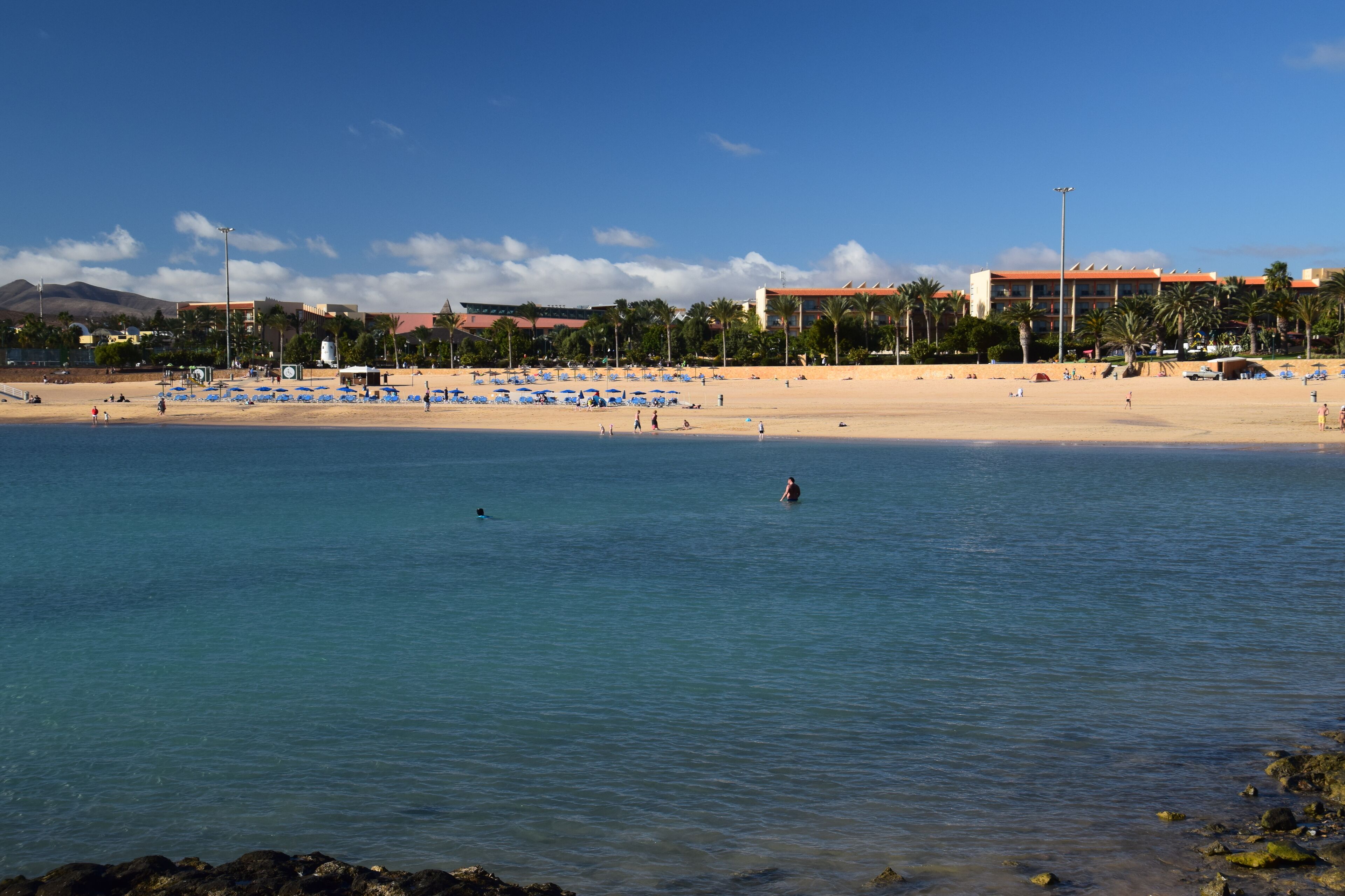 Una playa con agua azul cristalina, orilla de arena y un hotel al fondo.