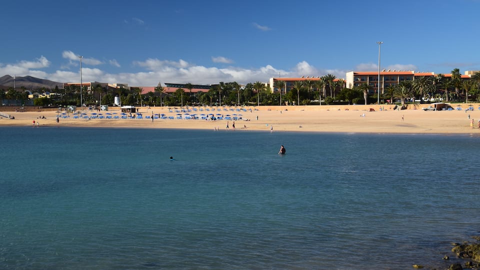Une plage avec une eau d’un bleu limpide, un rivage sablonneux et un hôtel en arrière-plan.