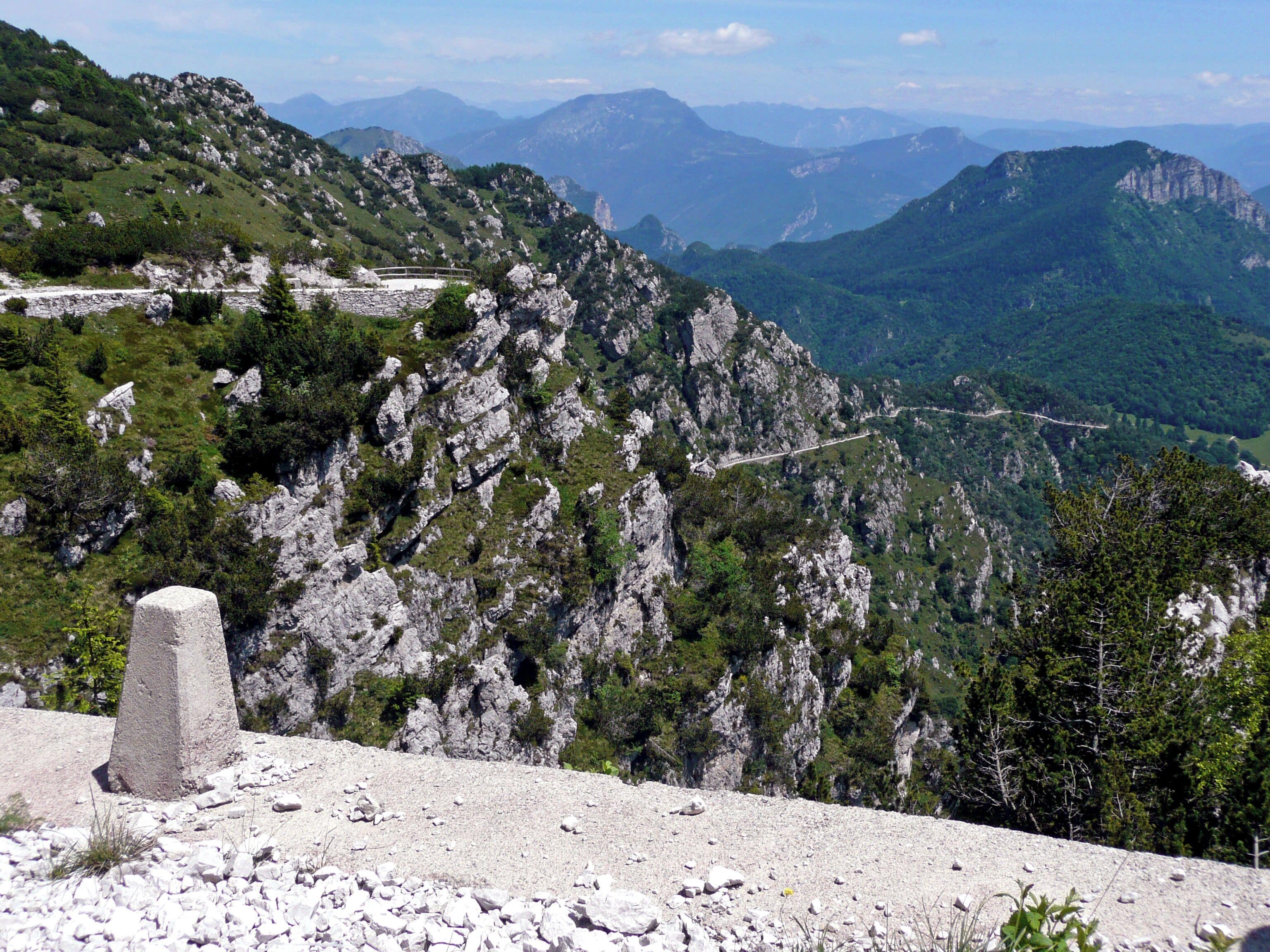 A rugged mountain landscape with rocky terrain, sparse vegetation, and distant mountain ranges under a clear sky.