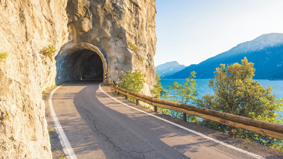 A winding road through a rocky gorge with a tunnel entrance, overlooking a lake and mountains.