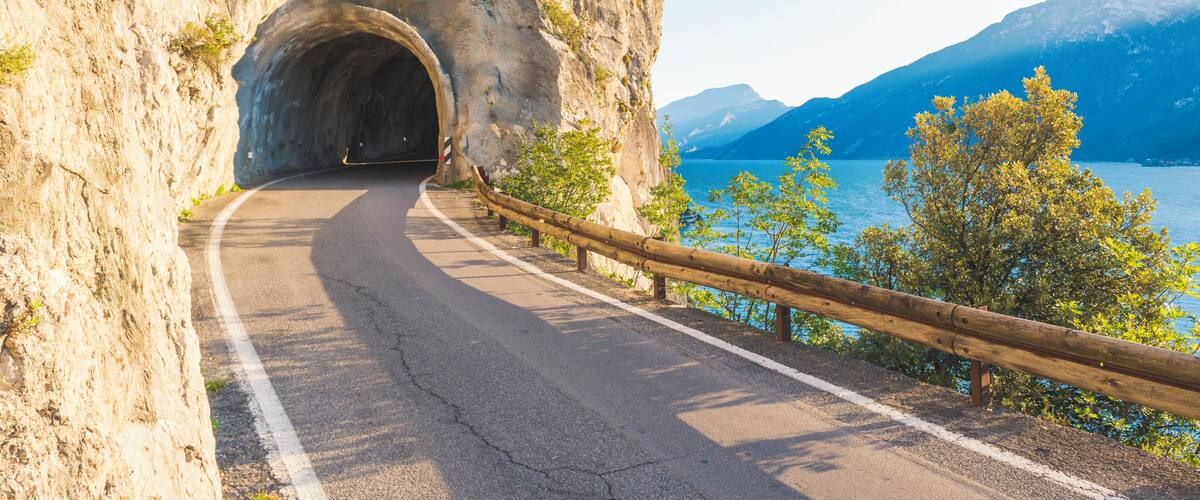 A winding road through a rocky gorge with a tunnel entrance, overlooking a lake and mountains.