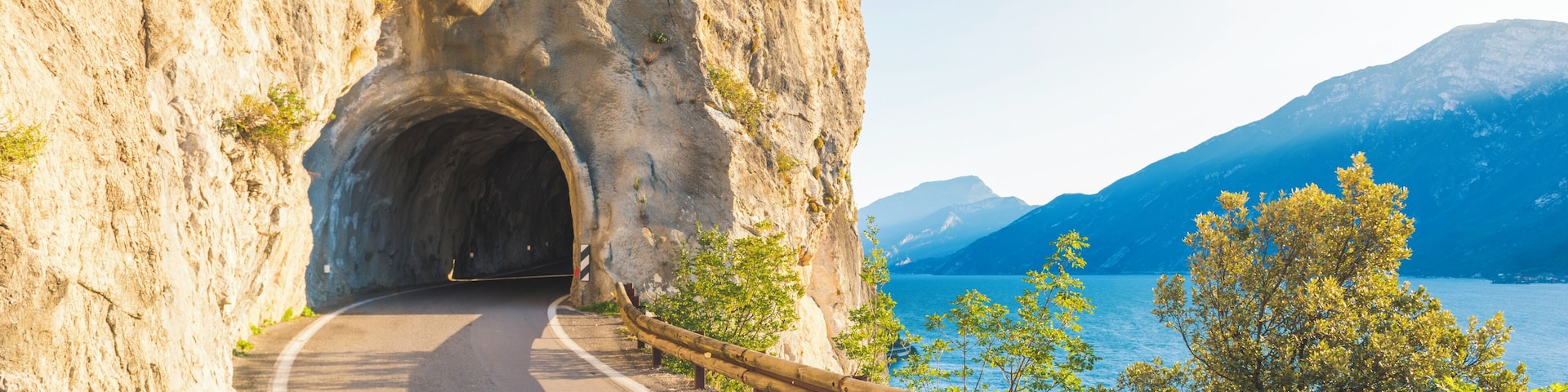 A winding road through a rocky gorge with a tunnel entrance, overlooking a lake and mountains.