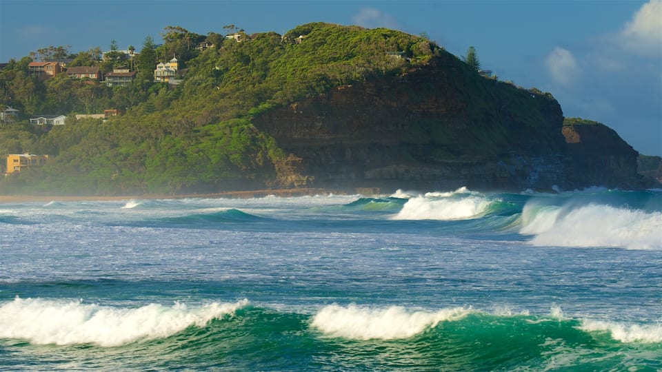 A coastal landscape with waves crashing onto a rocky shore, a cliff with houses, and lush greenery.