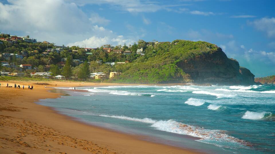 A beach with waves, a cliff, and houses on a hillside.