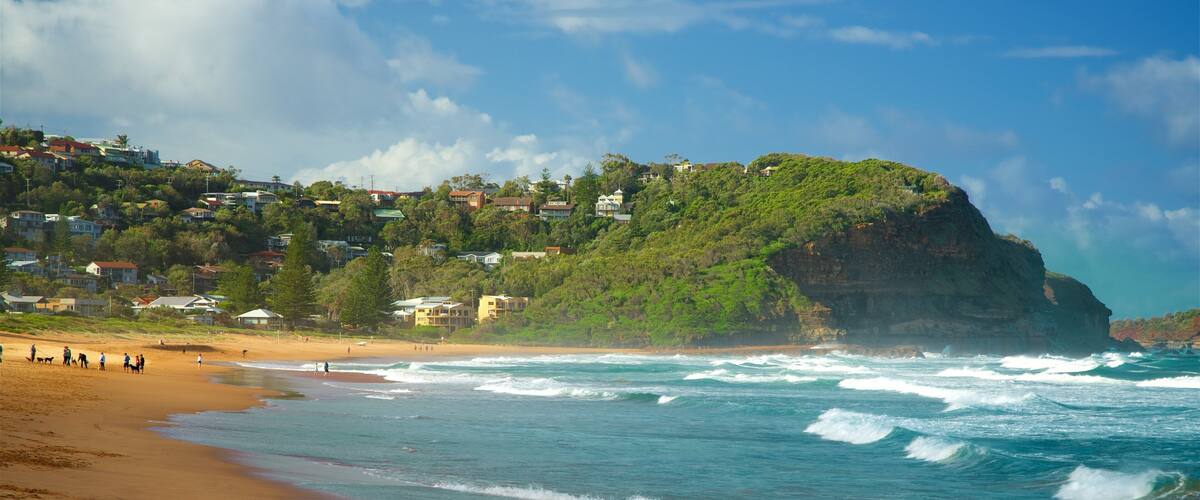 A beach with waves, a cliff, and houses on a hillside.