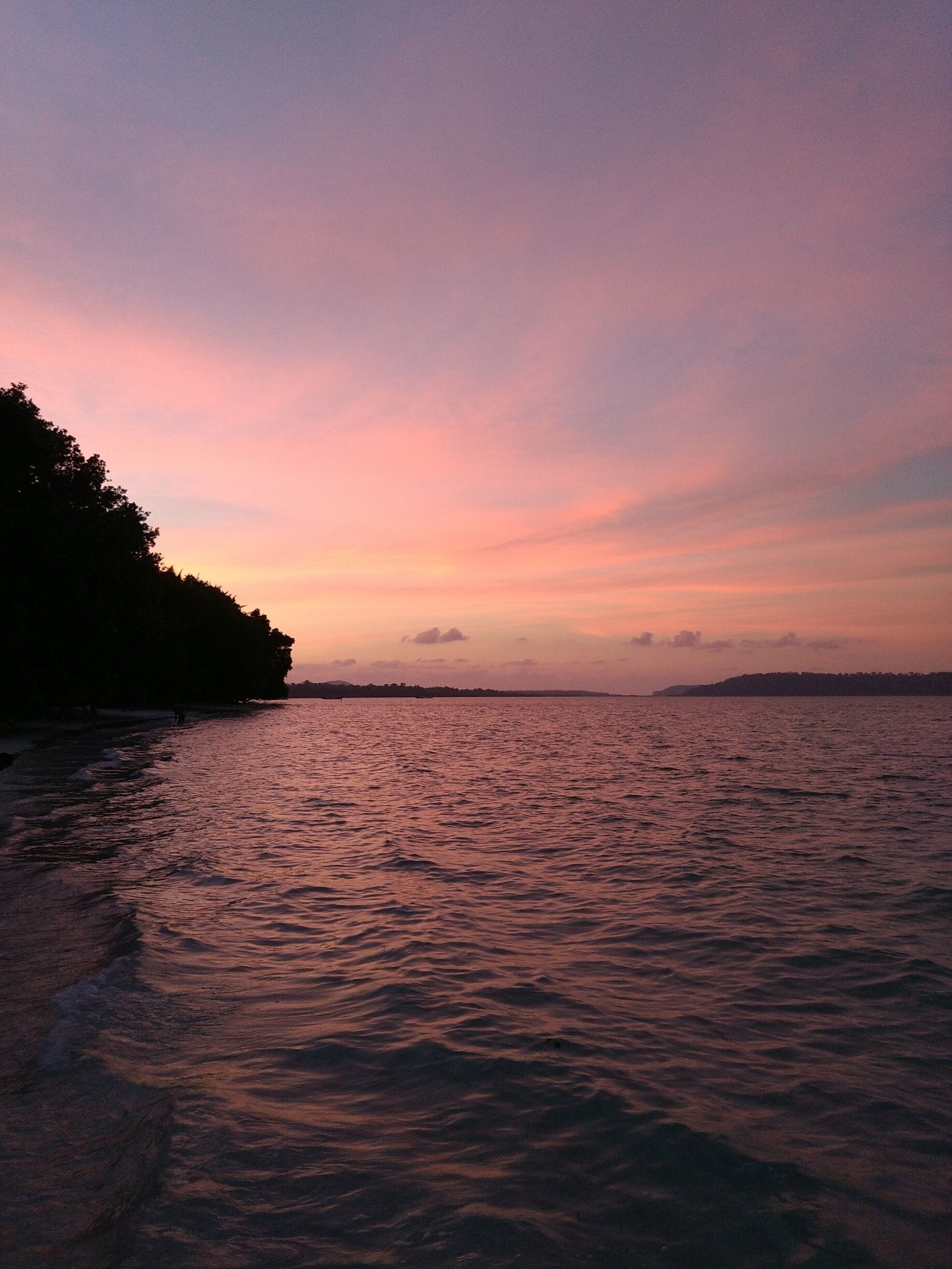 A sunset over a calm sea with a silhouette of a tree-covered landmass in the distance.