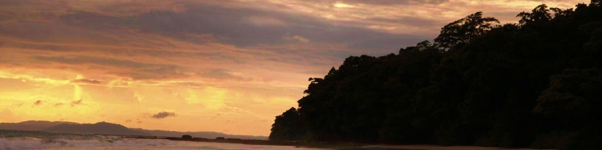 Una playa al atardecer con olas, un acantilado boscoso y una cadena montañosa distante.