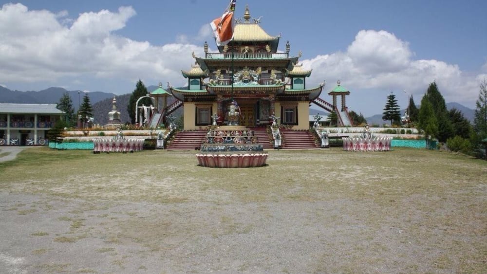 A traditional multi-tiered temple with a flag, surrounded by a grassy area and mountains in the background.