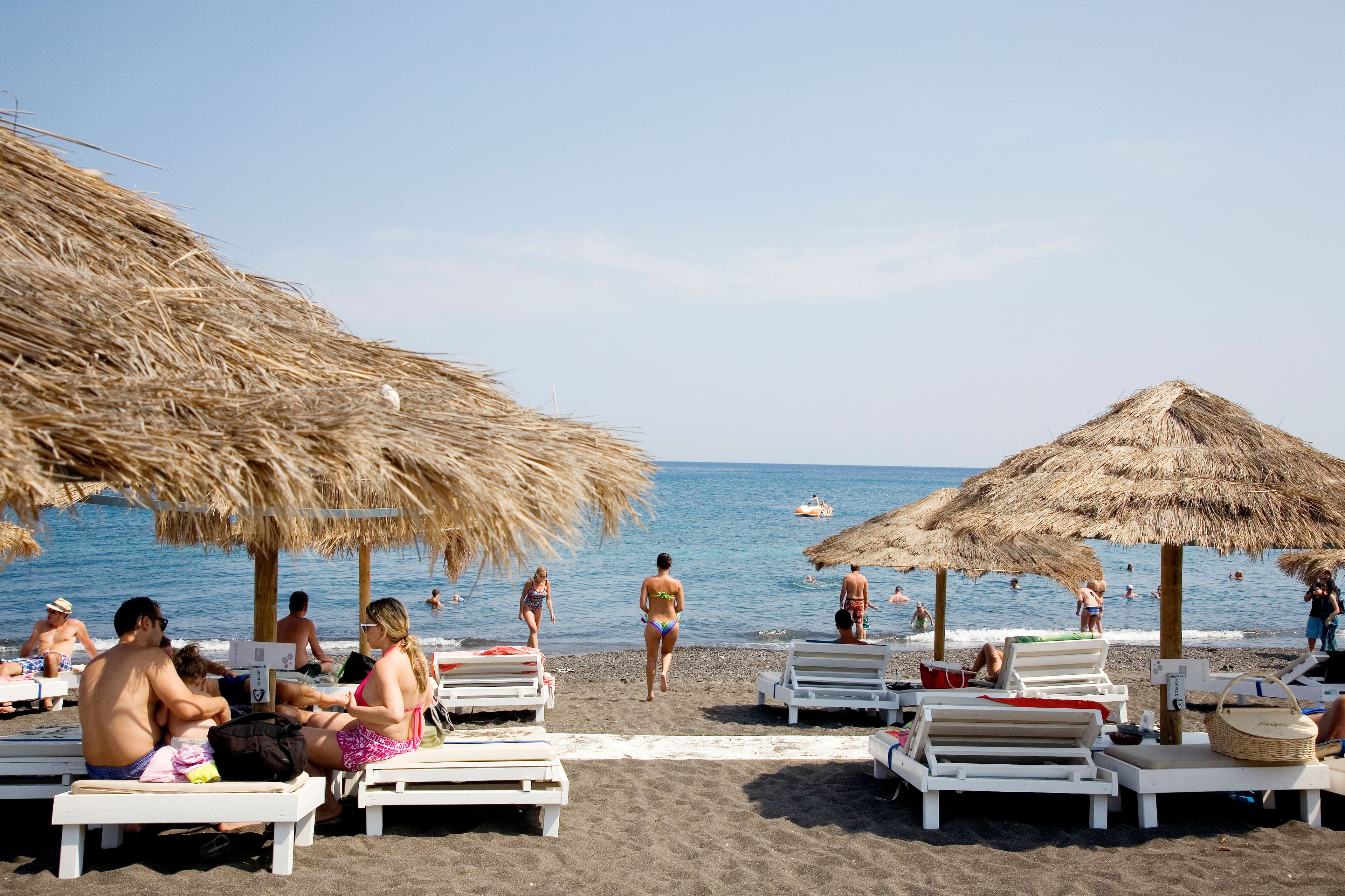 A beach with thatched umbrellas, sun loungers, and people swimming in the water.