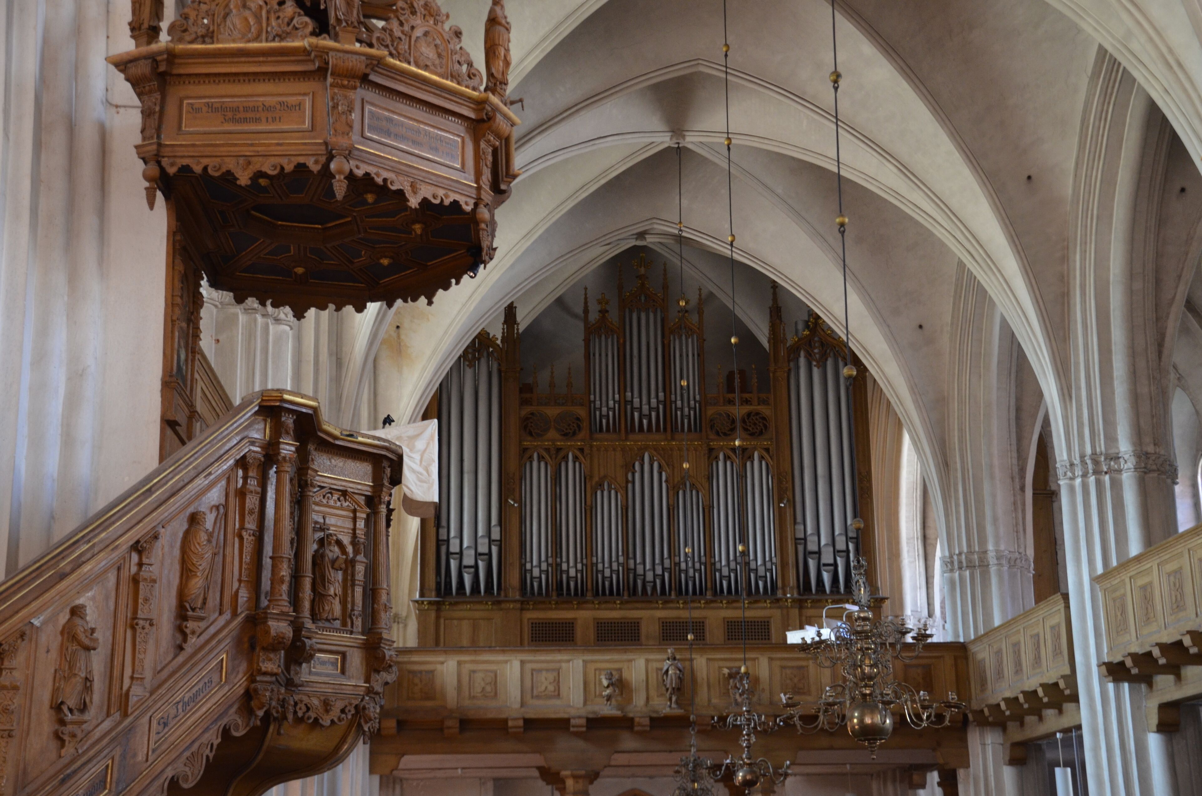 An ornate organ with multiple pipes, a wooden pulpit, and a chandelier in a cathedral.