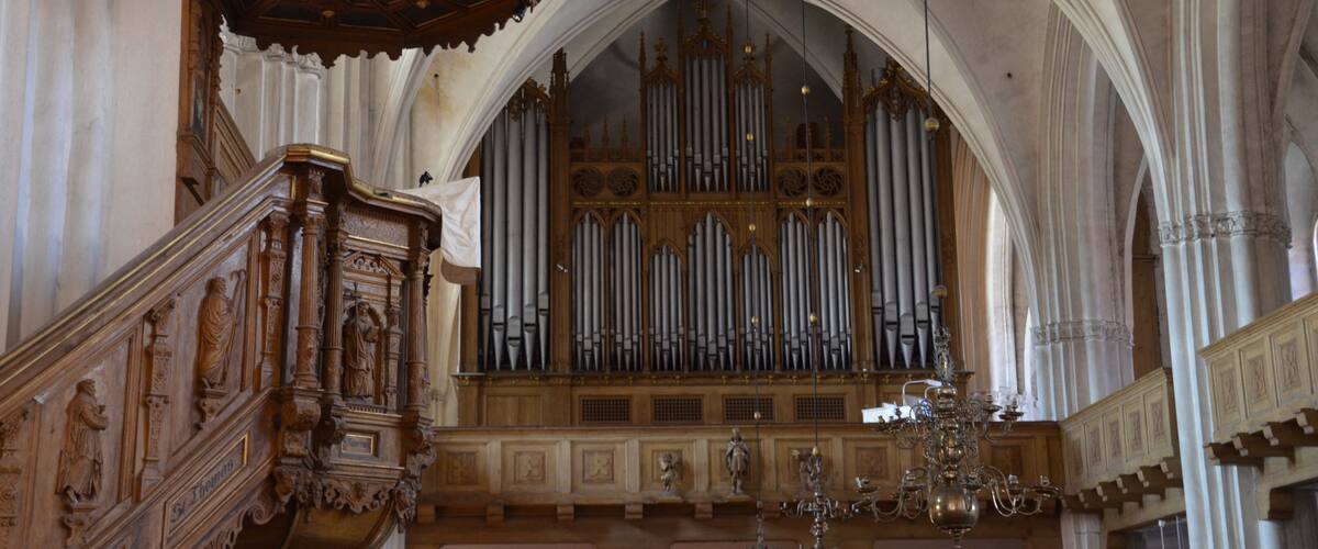An ornate organ with multiple pipes, a wooden pulpit, and a chandelier in a cathedral.