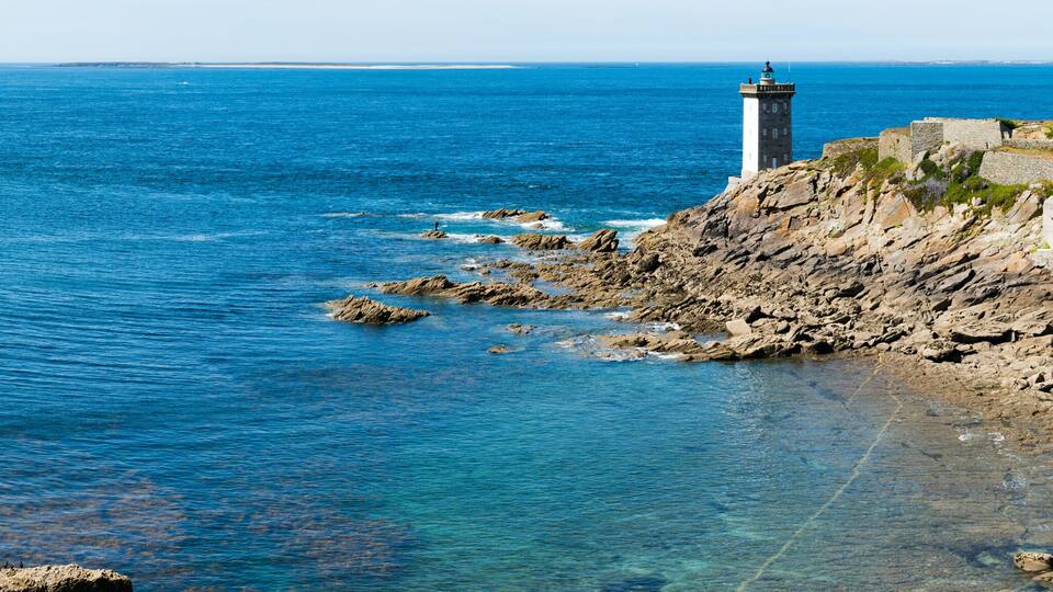 A coastal landscape with a lighthouse, rocky shoreline, and clear blue sea.