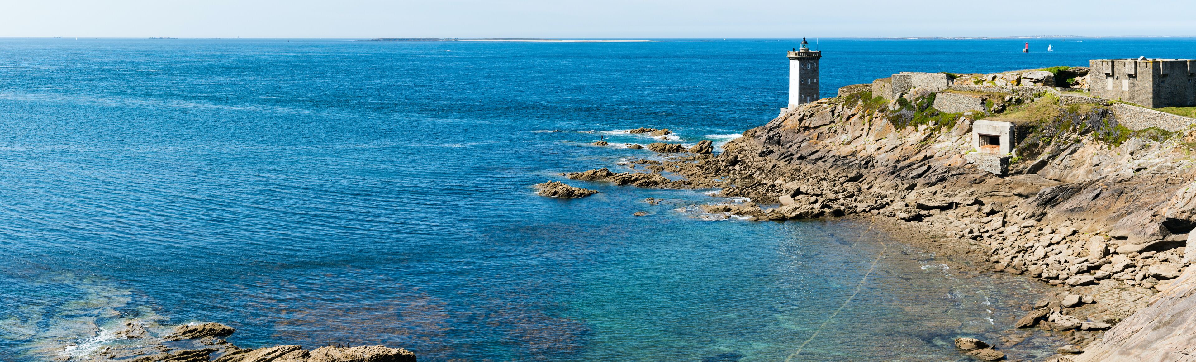 A coastal landscape with a lighthouse, rocky shoreline, and clear blue sea.