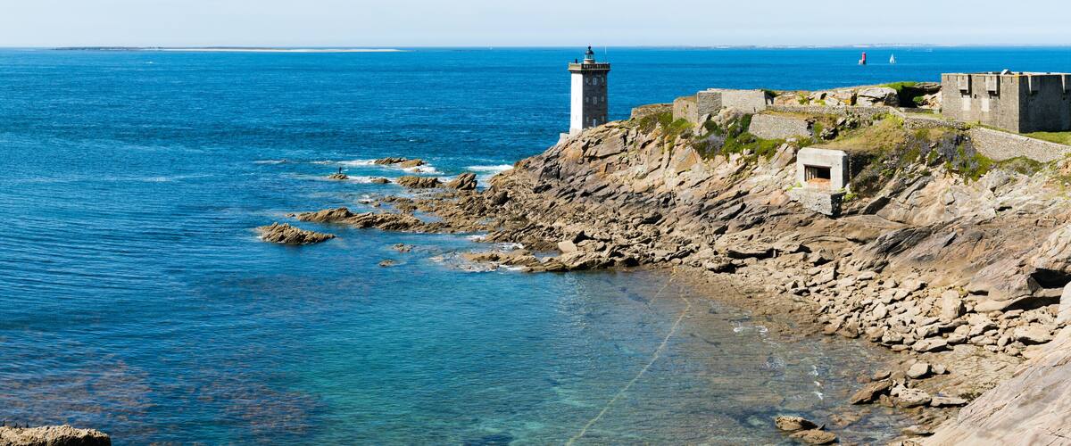 A coastal landscape with a lighthouse, rocky shoreline, and clear blue sea.