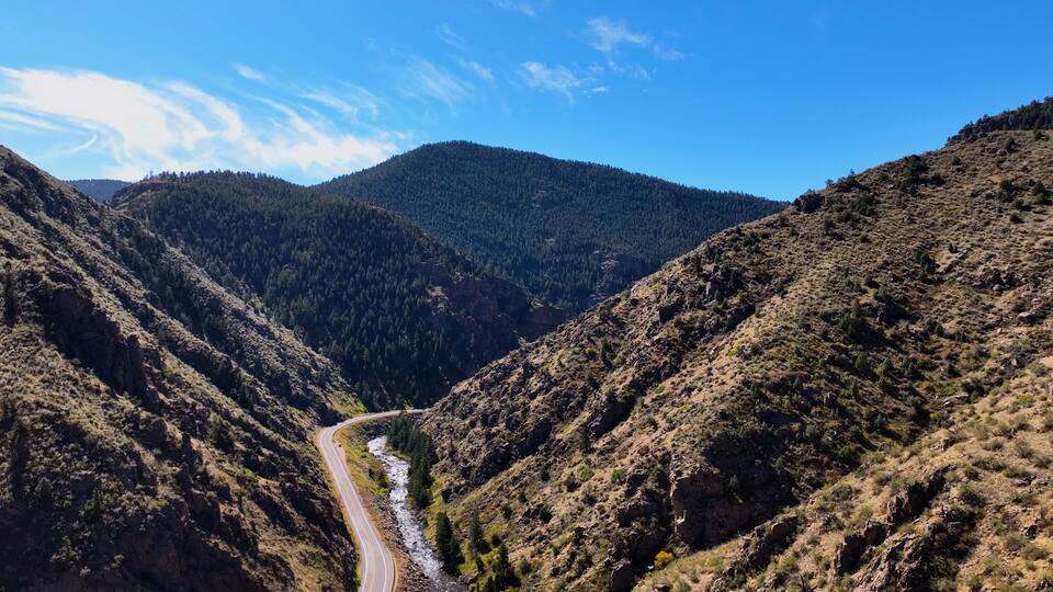 A winding road through a canyon with a river, surrounded by forested hills.