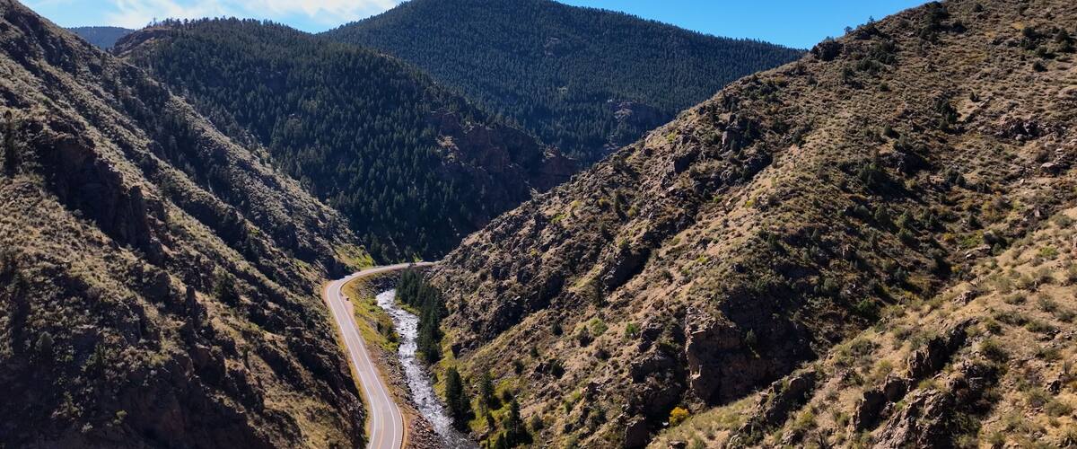 A winding road through a canyon with a river, surrounded by forested hills.