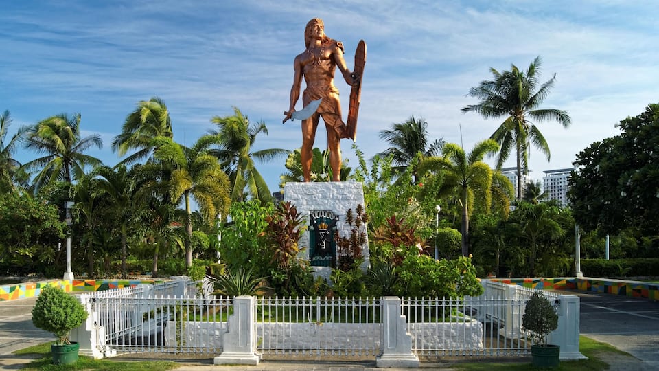 A large bronze statue of a warrior holding a sword and shield, surrounded by palm trees and a white fence.