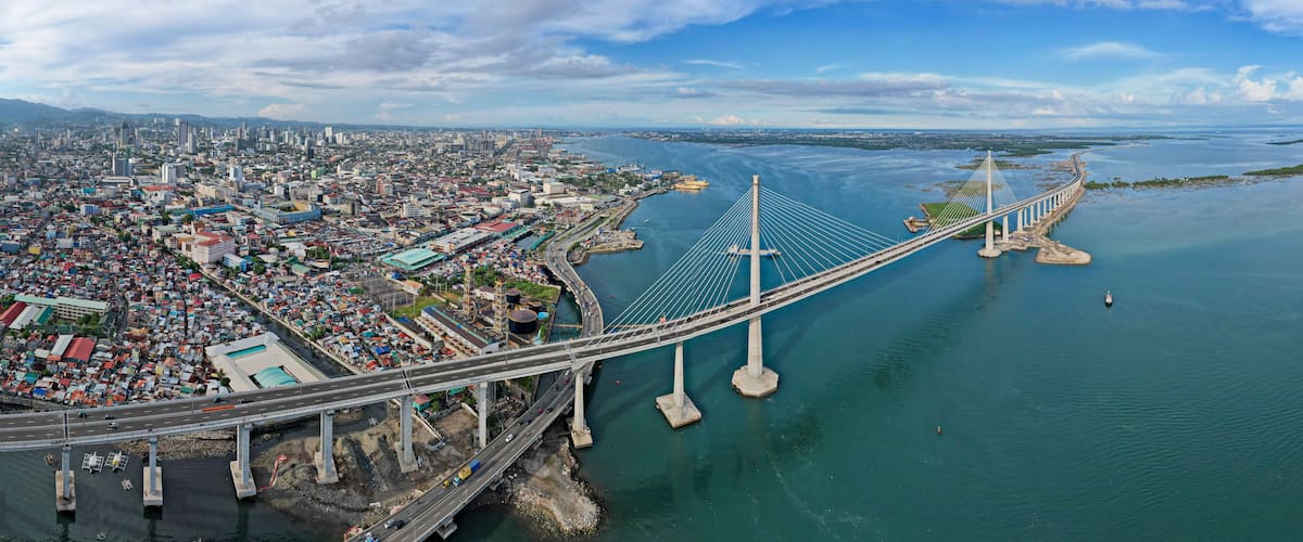 Un gran puente colgante cruza un río ancho, con una ciudad densamente poblada a un lado y aguas abiertas al otro.