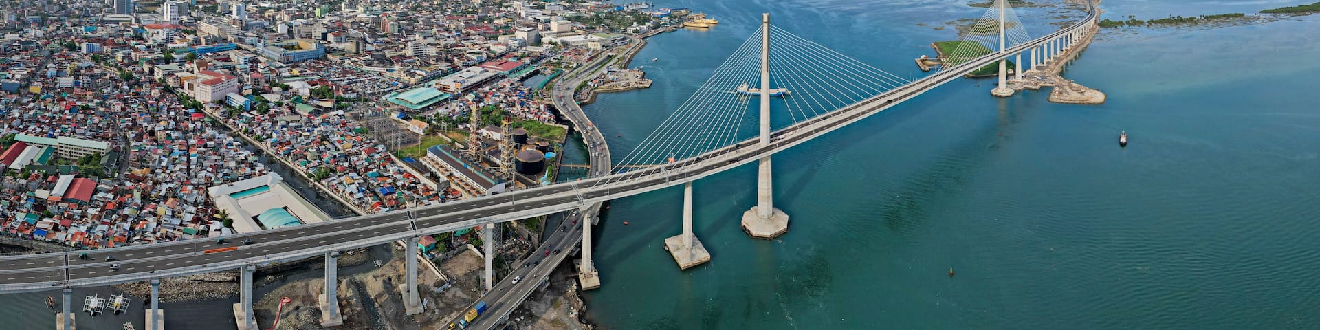 A large suspension bridge spanning a wide river, with a densely populated cityscape on one side and open water on the other.