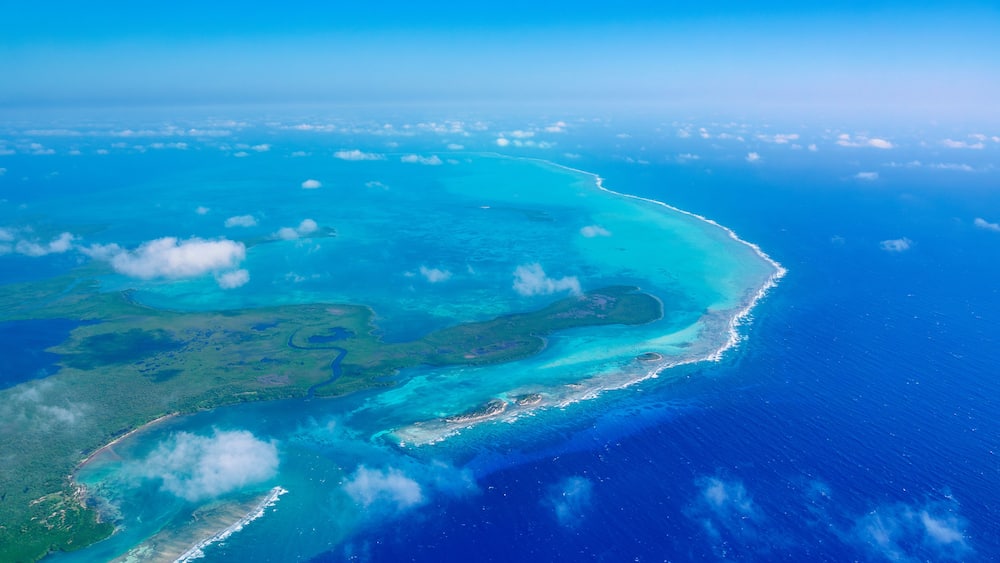 An aerial view of a coastal region with clear blue waters, a narrow beach, and scattered cloud formations.