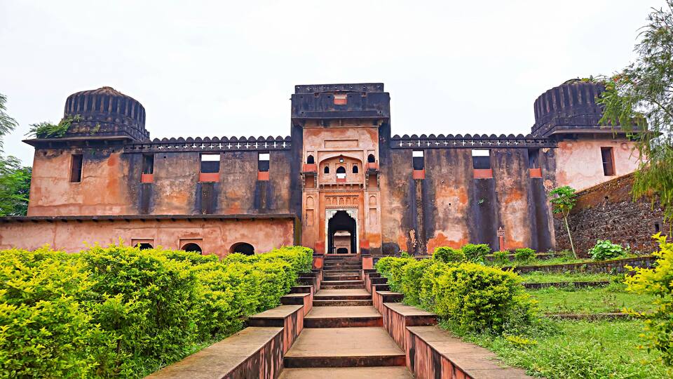 A large, historic building with a central archway, surrounded by lush greenery and a staircase leading up to it.