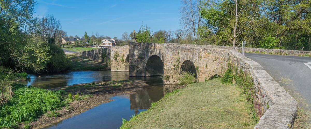 A stone bridge over a calm river, surrounded by greenery and trees.