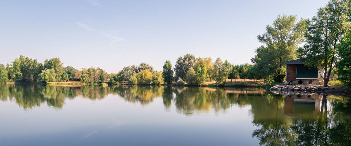 A calm lake with a wooden cabin on the right and trees reflecting on the water.