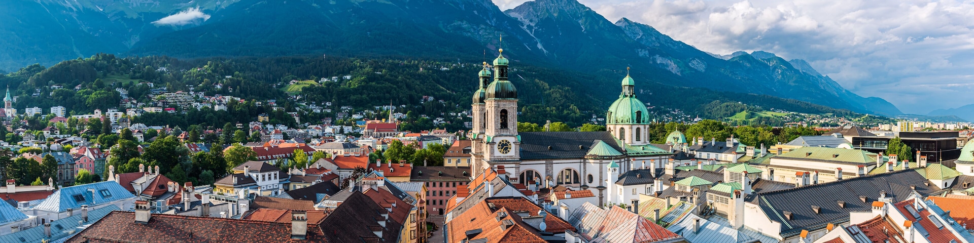 Uma cidade com edifícios históricos, uma igreja destacada com torre do relógio e montanhas ao fundo.