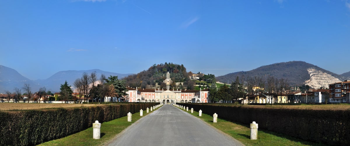 A road leading to a historic building with a tower, surrounded by mountains and residential houses.