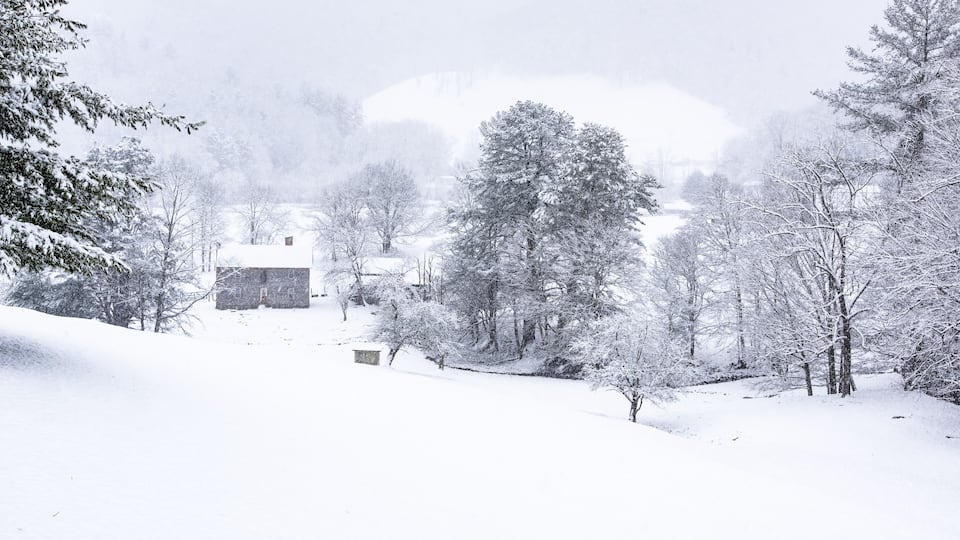 A snow-covered landscape with a stone building, trees, and a hill in the background.