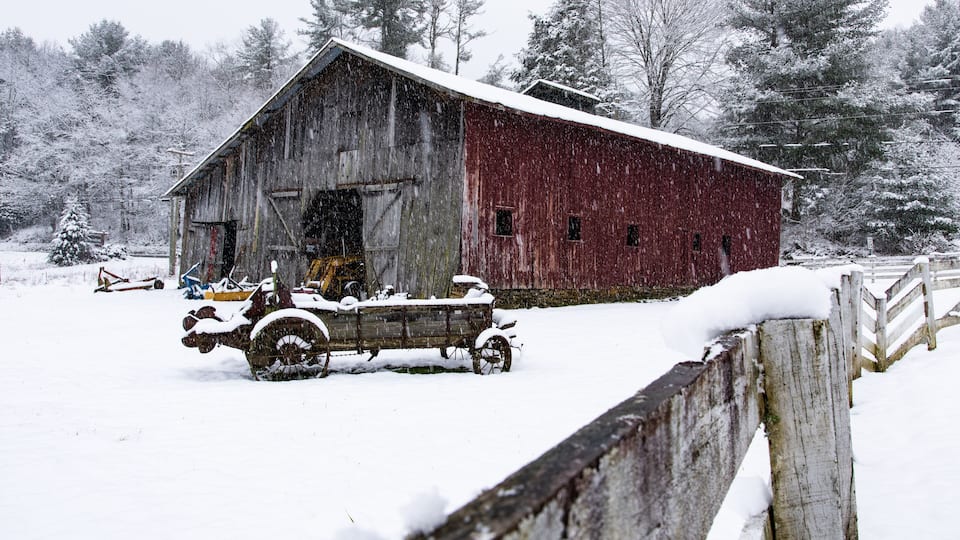 A snow-covered barn with a wooden fence in a snowy landscape.