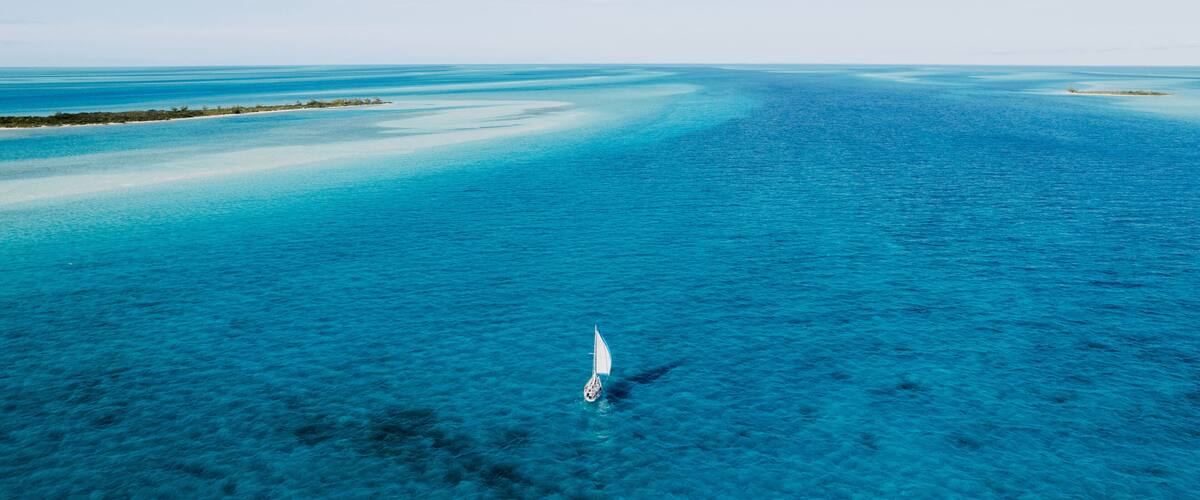 A clear blue sea with a sailboat in the distance and a narrow sandbar on the left.
