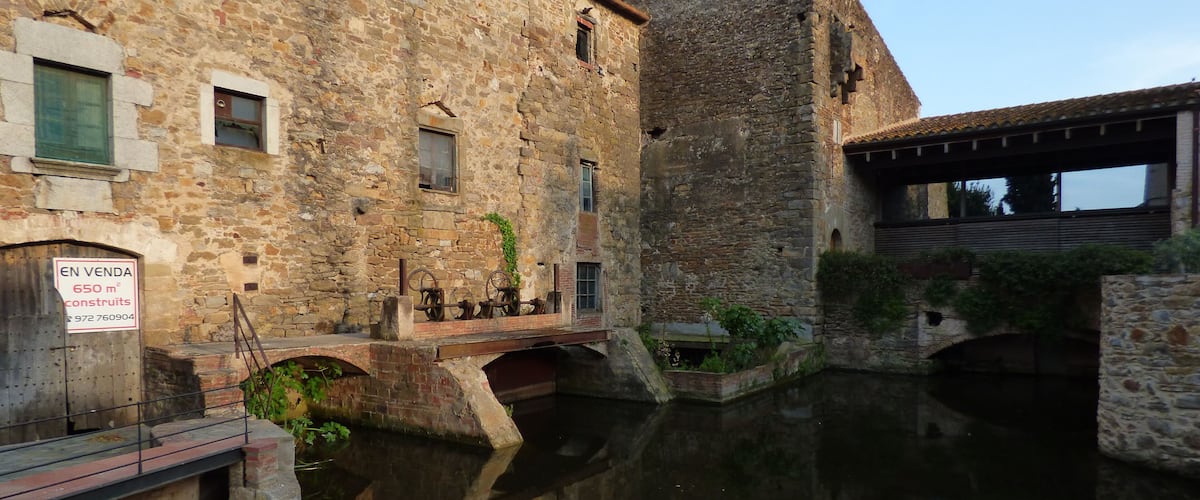 A stone building with a sign indicating it is for sale, a small bridge over a canal, and a reflection in the water.
