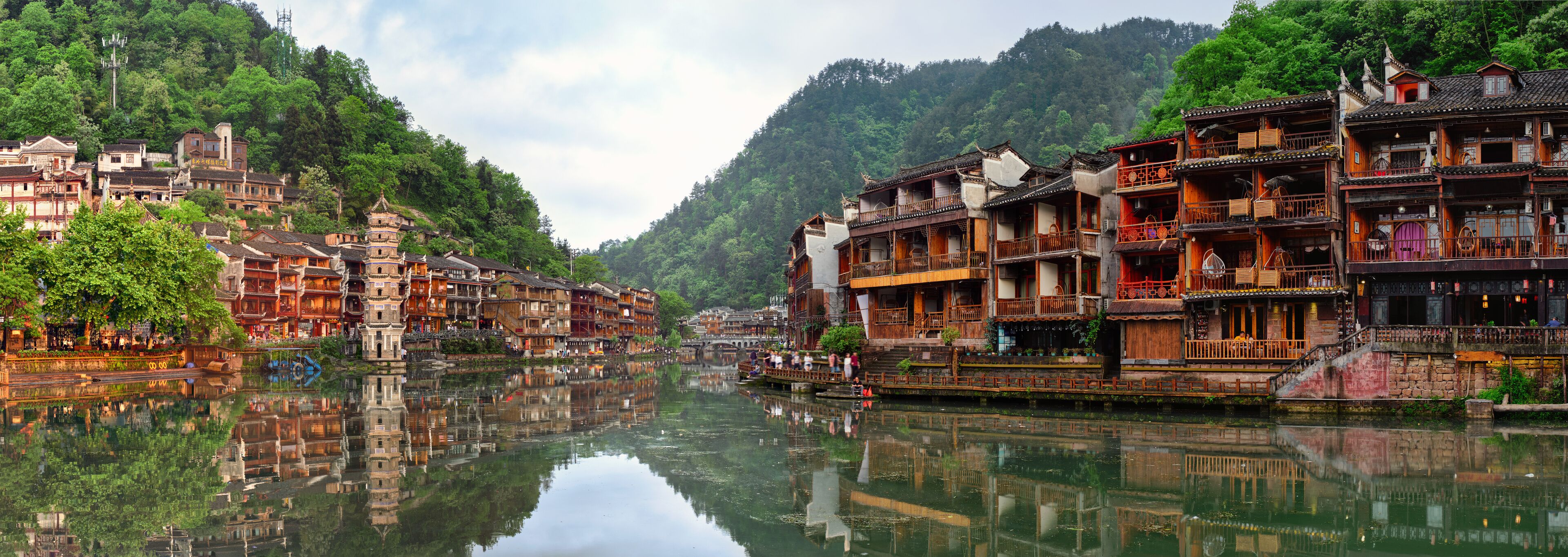A traditional riverside village with wooden buildings, a clear river, and lush green hills in the background.