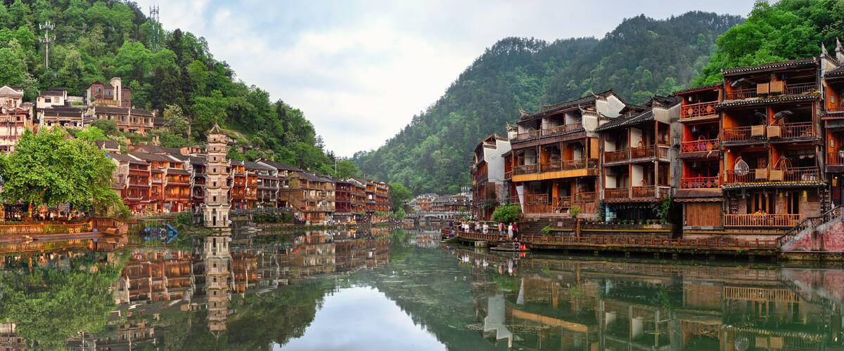 A traditional riverside village with wooden buildings, a clear river, and lush green hills in the background.