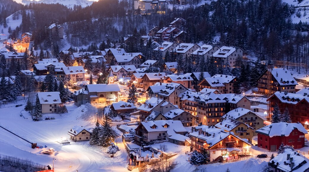 Aerial view of a snow-laden village nestling beneath majestic mountains, lights twinkling like stars against the cold, twilight sky, Claviere, Piedmont, Italy.