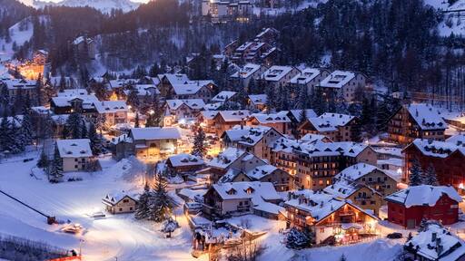 Aerial view of a snow-laden village nestling beneath majestic mountains, lights twinkling like stars against the cold, twilight sky, Claviere, Piedmont, Italy.
