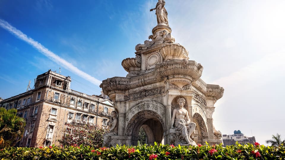 A classical fountain with statues, surrounded by lush greenery and a historic building in the background.